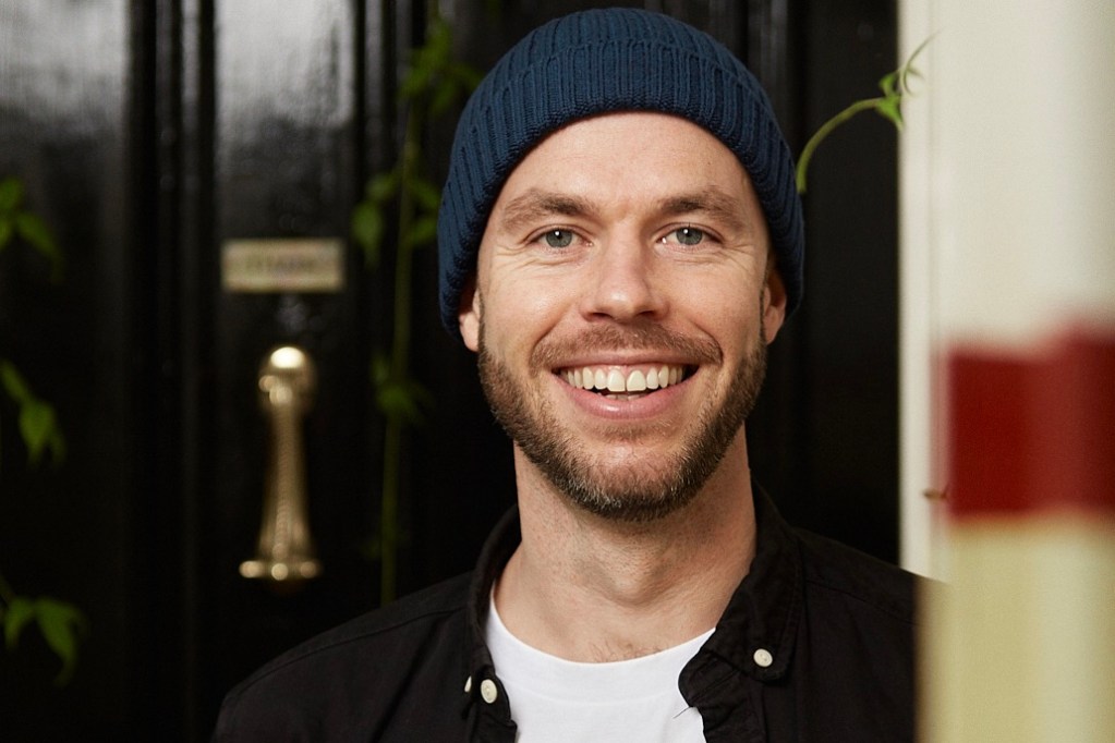 Smiling man wearing a blue beanie, standing in front of a dark door with a brass handle, surrounded by greenery.
