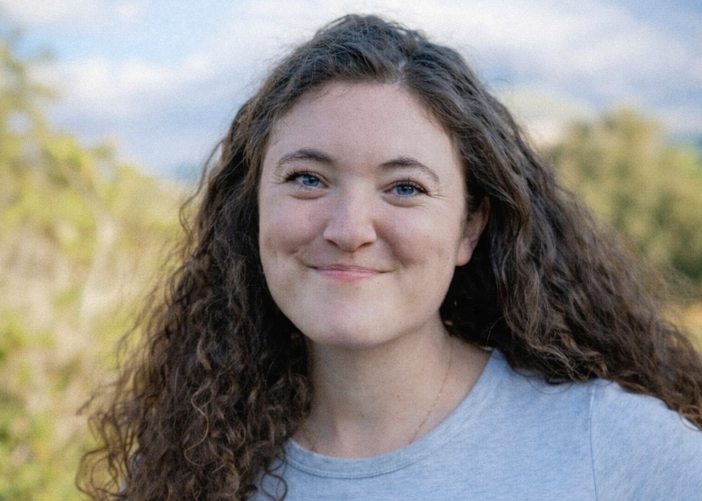 A smiling woman with curly hair and blue eyes, wearing a gray t-shirt, stands outdoors with a blurred natural background.