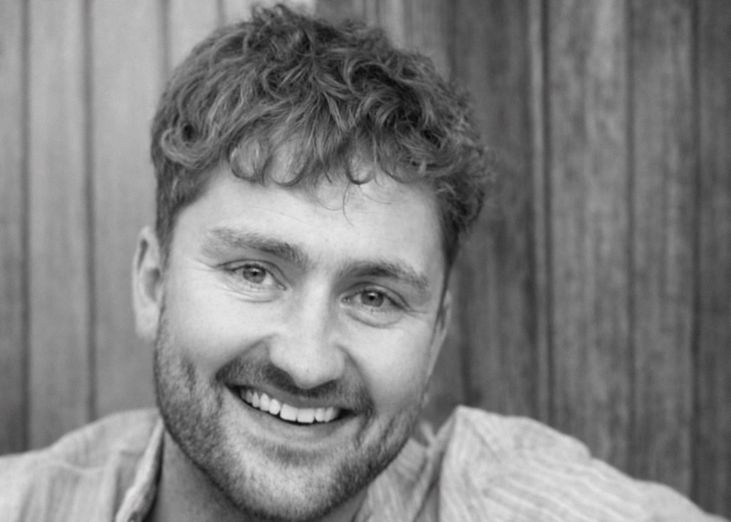 A smiling man with curly hair and a beard, sitting in front of a wooden background.