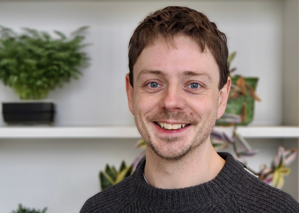 Smiling man with short hair wearing a dark sweater, standing in front of a shelf with various green plants.
